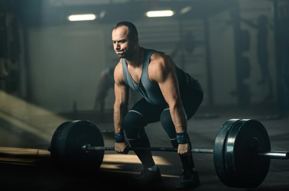 A man in a tank top lifting a heavy barbell in a dimly lit gym