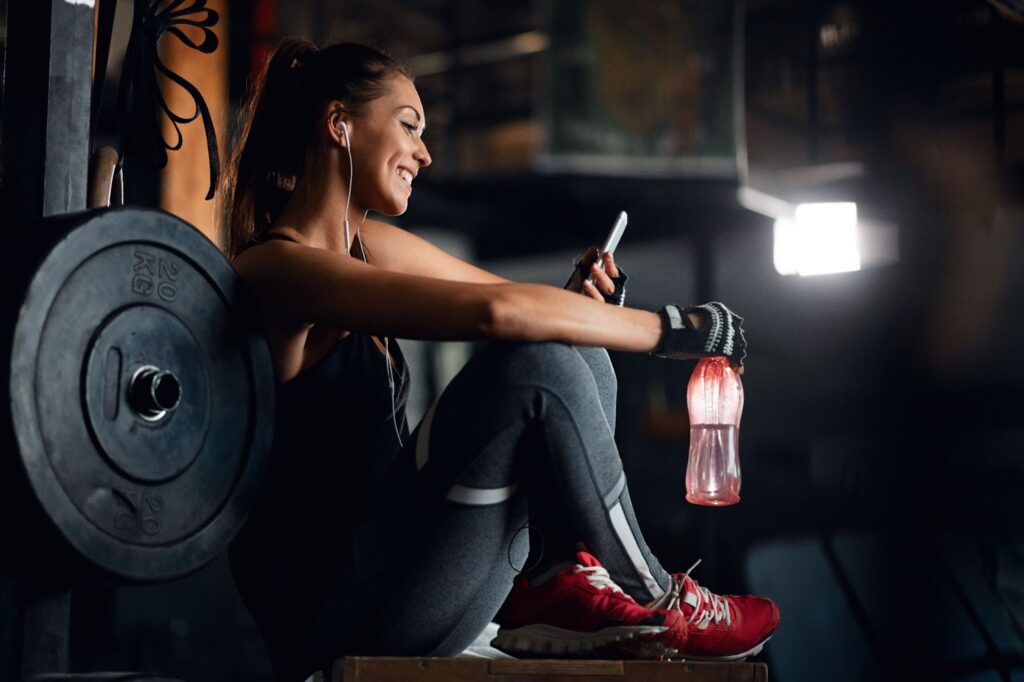 Smiling woman relaxing on water break and reading text message on her phone in gym